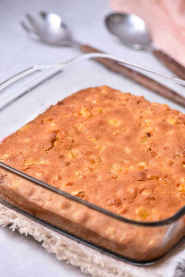 A square glass baking dish holds a freshly baked cornbread with a golden-brown crust, reminiscent of homemade corn pudding without Jiffy mix. The dish is placed on a tan woven trivet, and two silver spoons are in the background on a light-colored surface.