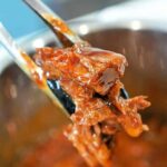 Tongs holding shredded instant pot BBQ beef with sauce, above a pot of more saucy beef.