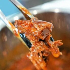 Tongs holding shredded instant pot BBQ beef with sauce, above a pot of more saucy beef.