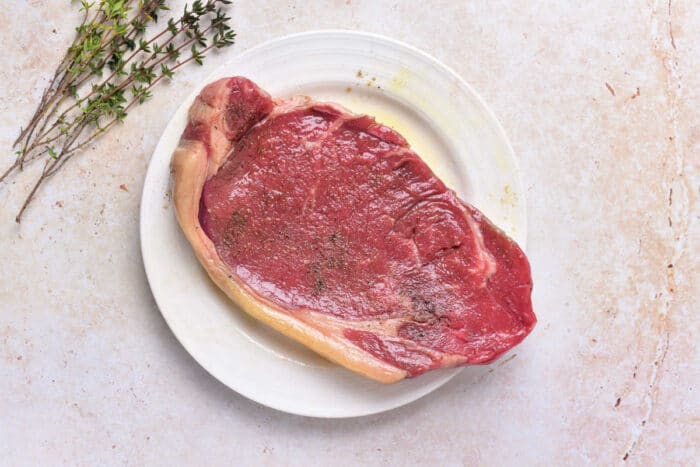 Raw ribeye steak sits on a white plate, seasoned with a touch of pepper and salt. Fresh thyme sprigs are placed next to the plate on a light-colored surface. For tips on preparing similar cuts, see how to cook beef strip loin steaks.