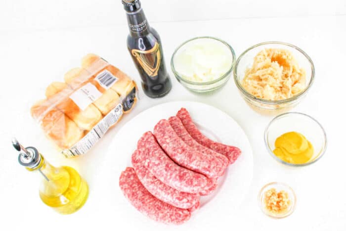 A variety of ingredients are laid out on a white surface for how to cook bratwurst. Included are six raw sausages on a white plate, a bottle of Guinness beer, a pack of Hawaiian rolls, a bottle of olive oil, and bowls containing sliced onions, sauerkraut, mustard, and minced garlic.