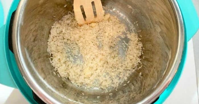 Uncooked rice being stirred with a wooden spatula in a stainless steel pot on a teal base, ready to become a delicious pressure cooker coconut rice pudding.