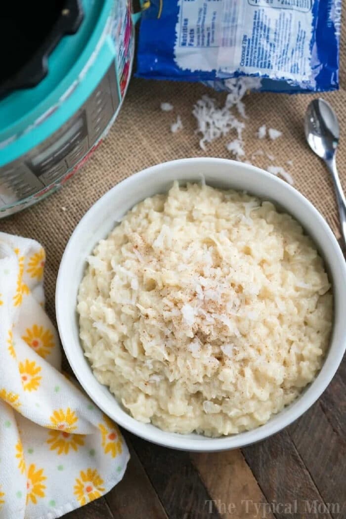 A bowl of creamy pressure cooker coconut rice pudding, topped with shredded coconut, sits beside a spoon and a yellow floral napkin.