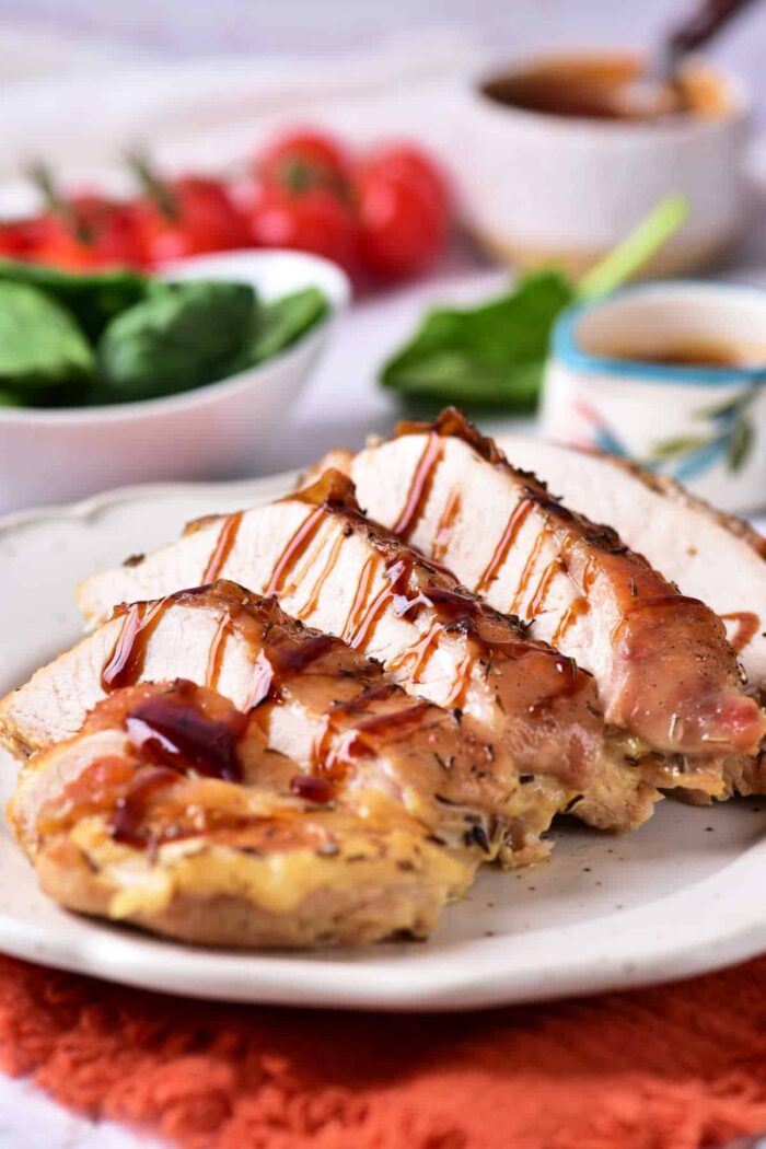 A plate with sliced roasted chicken breast drizzled with a dark sauce, hinting at the richness of Turkey Confit. A side dish, possibly a small bowl of green leafy vegetables, and a sauce bowl are in the background.