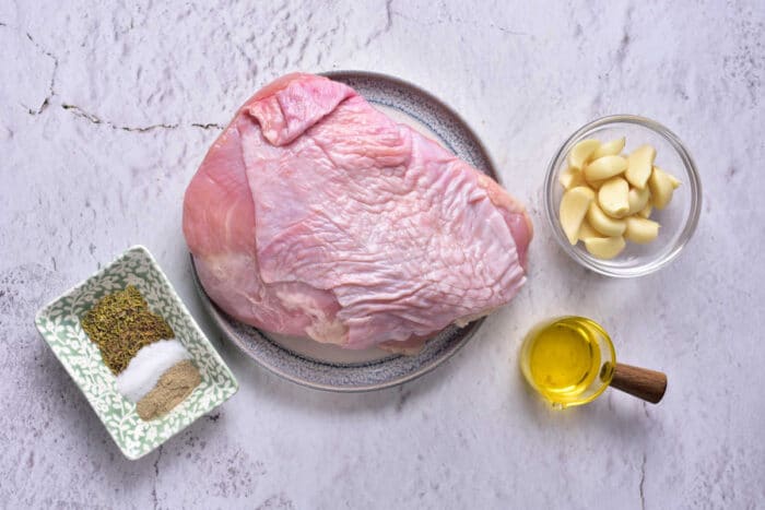 A raw turkey breast on a plate is placed on a kitchen countertop. Surrounding it are a small dish of spices, including salt and pepper, a bowl with peeled garlic cloves, and a small container of olive oil—perfect ingredients for preparing Turkey Confit.