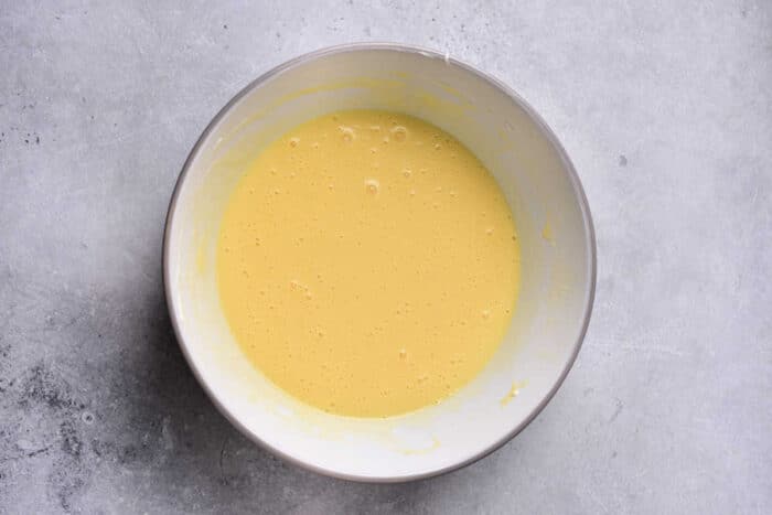 A white bowl filled with a light yellow, smooth, and slightly bubbly mixture is placed on a gray, textured surface. The mixture appears to be a homemade batter for Corn Pudding Without Jiffy Mix.