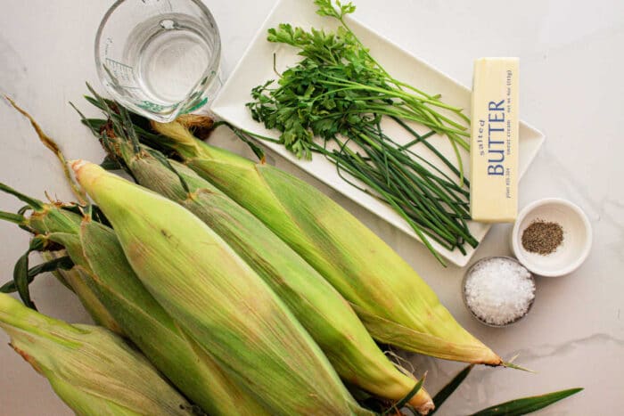 An arrangement for Crockpot Corn on the Cob: fresh corn on the cob, a glass of water, a bunch of parsley and chives on a white plate, unsalted butter, and small bowls of salt and pepper on a pristine white surface.