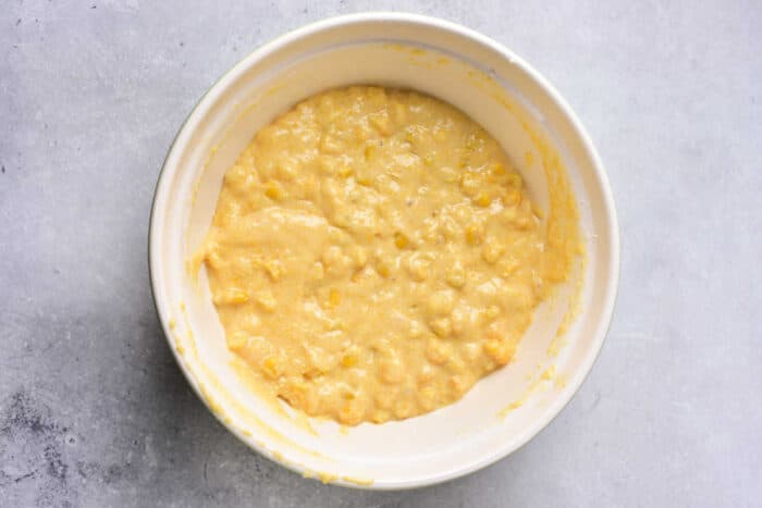 A white bowl filled with thick, yellowish batter containing visible corn kernels, placed on a light gray surface—perfect for homemade Corn Pudding Without Jiffy Mix.