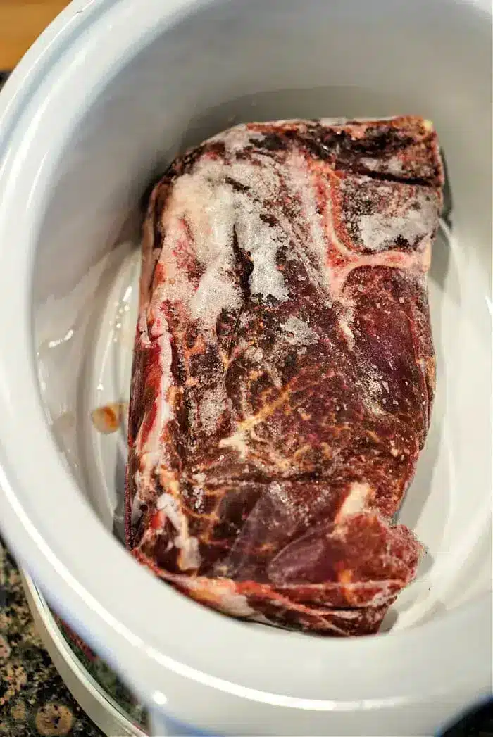 A frozen beef chuck roast rests in a slow cooker, marbled with fat and covered in frost. It sits elegantly in a white ceramic dish on a granite countertop, promising tender perfection.