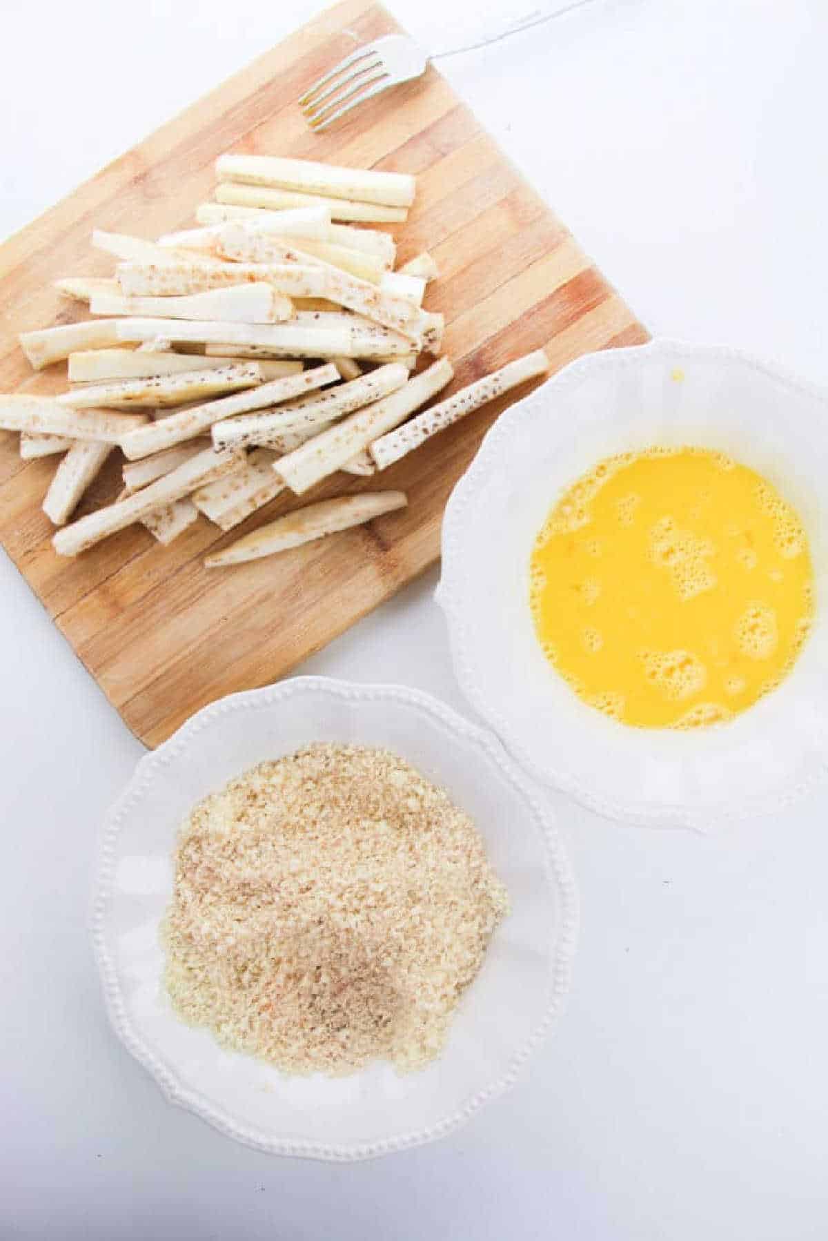 eggplant sticks on a cutting board, with bowls of beaten eggs and breadcrumbs next to them&mdash;perfect for making unique alternatives to Eggplant Fries.