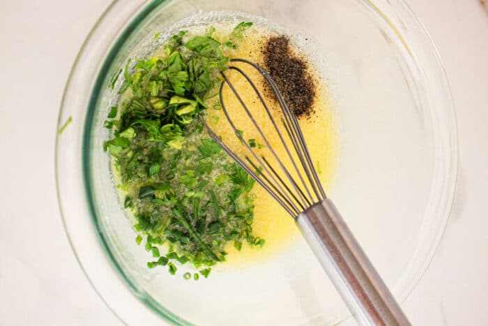 A glass mixing bowl containing a yellow liquid with finely chopped green herbs, ground black pepper, and a metal whisk, reminiscent of seasonings for Crockpot Corn on the Cob. The bowl is placed on a light-colored surface.