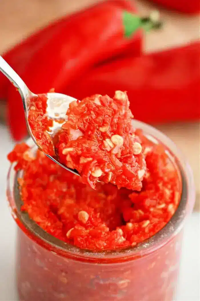 A spoonful of bright red garlic chili paste is lifted from a jar. The paste appears chunky with visible seeds, hinting at its rich texture. In the background, whole red peppers are partially visible on a wooden surface, inspiring culinary creativity with this vibrant recipe.