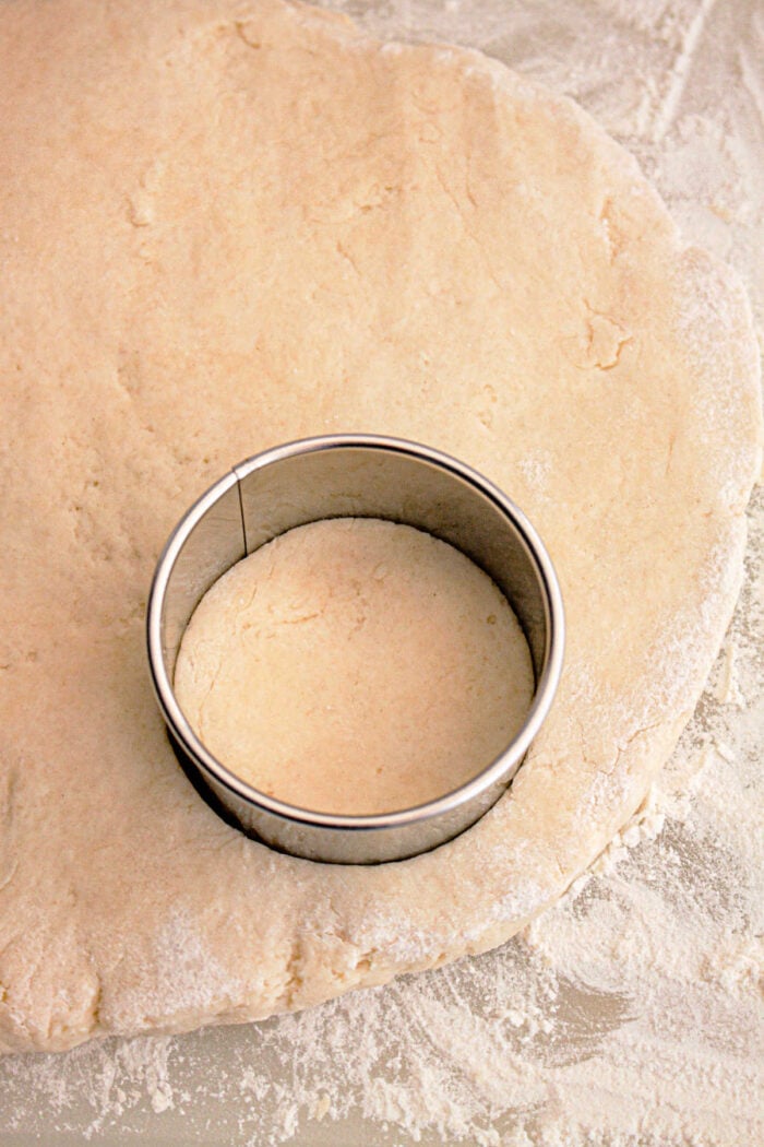 A metal round cookie cutter is pressed into rolled-out dough on a floured surface, creating a perfect shape for Slow Cooker Biscuits and Gravy.