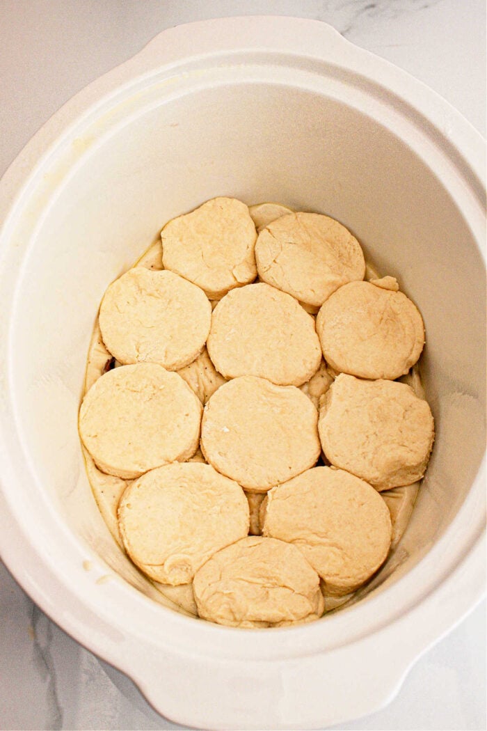 A white slow cooker with a lid open, showcasing an arrangement of what appears to be raw dough biscuits neatly placed inside. The interior and the sides of the slow cooker are visible. The background shows a grayish-white surface, setting the stage for some delicious Slow Cooker Biscuits and Gravy.