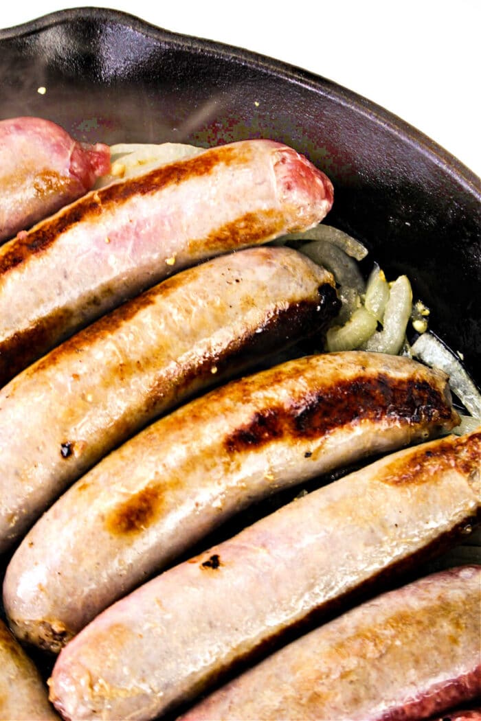 Five sausages are cooking in a cast iron skillet. The sausages have a browned, crispy exterior and are lying on a bed of sliced onions. The image shows the pan from an overhead perspective, offering a delectable hint on how to cook bratwurst to perfection.