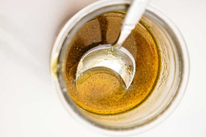 Close-up view of a glass jar filled with a golden-brown liquid, likely a sauce or dressing perfect for slow cooker vegetables, with a metal spoon partially submerged in it. The liquid has some visible seasoning or spices. The background is white and minimal.