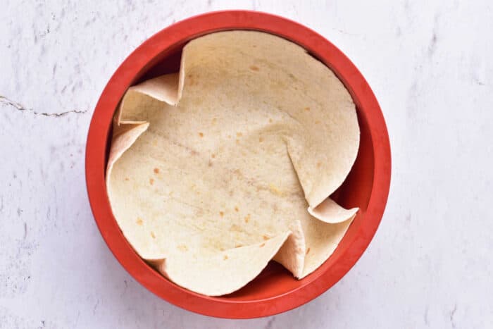 A single tortilla is arranged inside a red, round baking dish on a light-colored surface. The edges are slightly folded and overlapping, forming a semi-crumpled appearance, perfect for a delicious cottage cheese quiche. The background is textured with subtle cracks and lines.