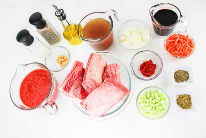 A collection of ingredients for a recipe arranged on a white surface, including raw beef short ribs perfect for Short Rib Tacos, tomato sauce, beef broth, diced onions, shredded carrots, chopped celery, red wine, tomato paste, minced garlic, cooking oil, salt, pepper and dried herbs.