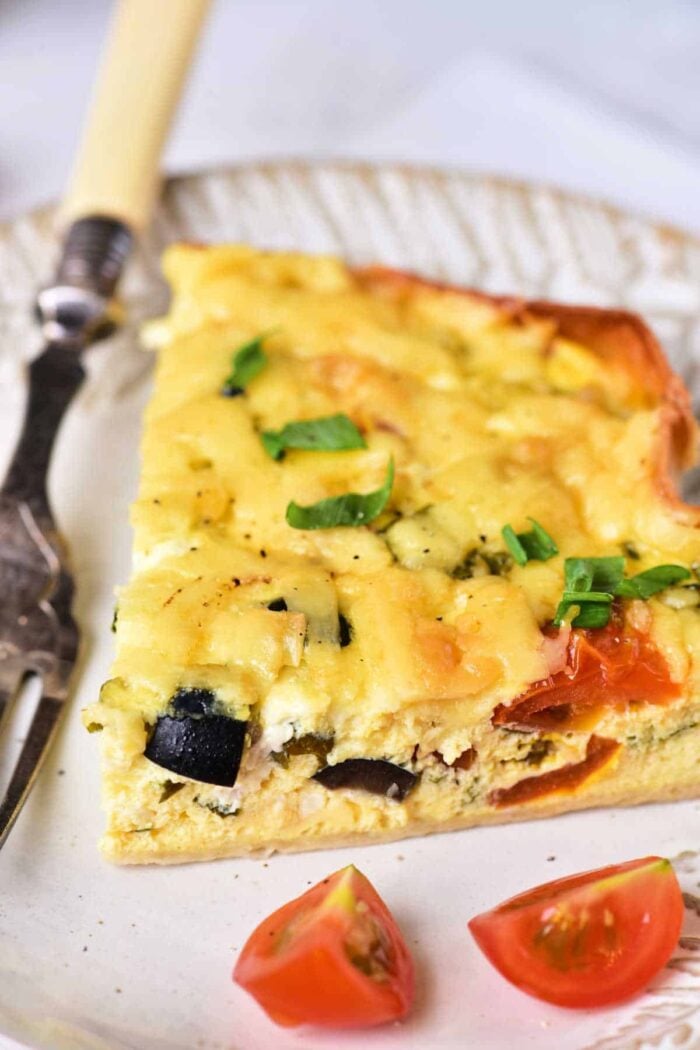 A slice of vegetable cottage cheese quiche on a plate. The quiche has visible pieces of zucchini, tomato, and a topping of melted cheese, garnished with chopped green onions. A fork with a decorative handle is placed next to the quiche. Two cherry tomato halves are on the plate.