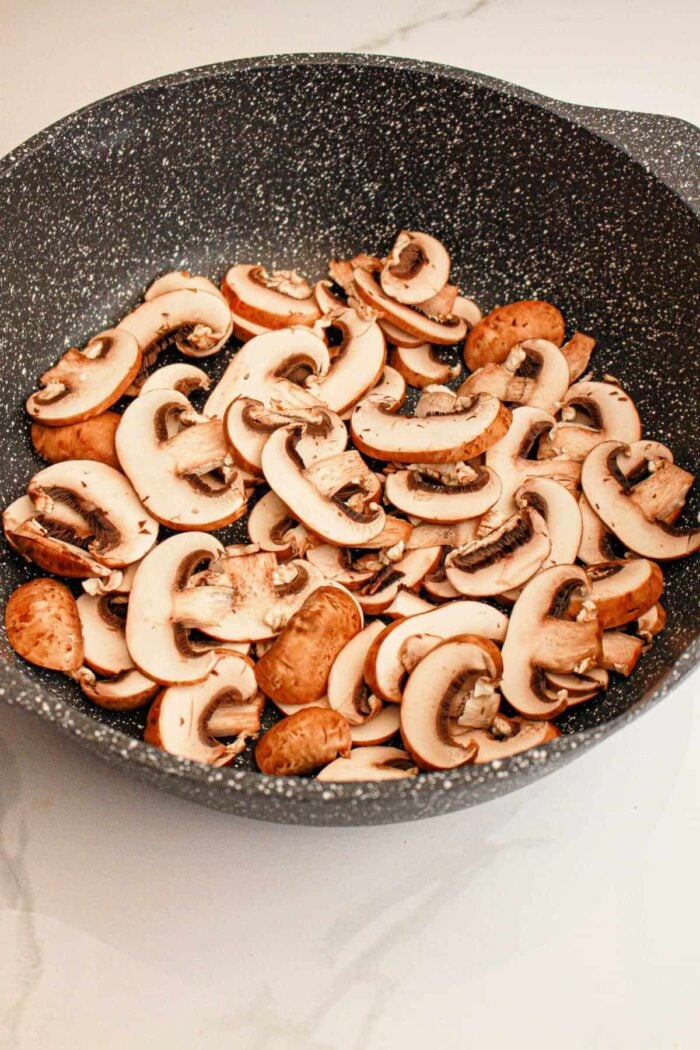 A black, speckled frying pan filled with slices of raw mushrooms sits invitingly on a light-colored countertop, setting the perfect stage for a hearty Slow Cooker Biscuits and Gravy breakfast.