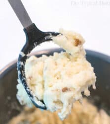 A potato masher holding a scoop of creamy slow cooker mashed potatoes with potato skin pieces.