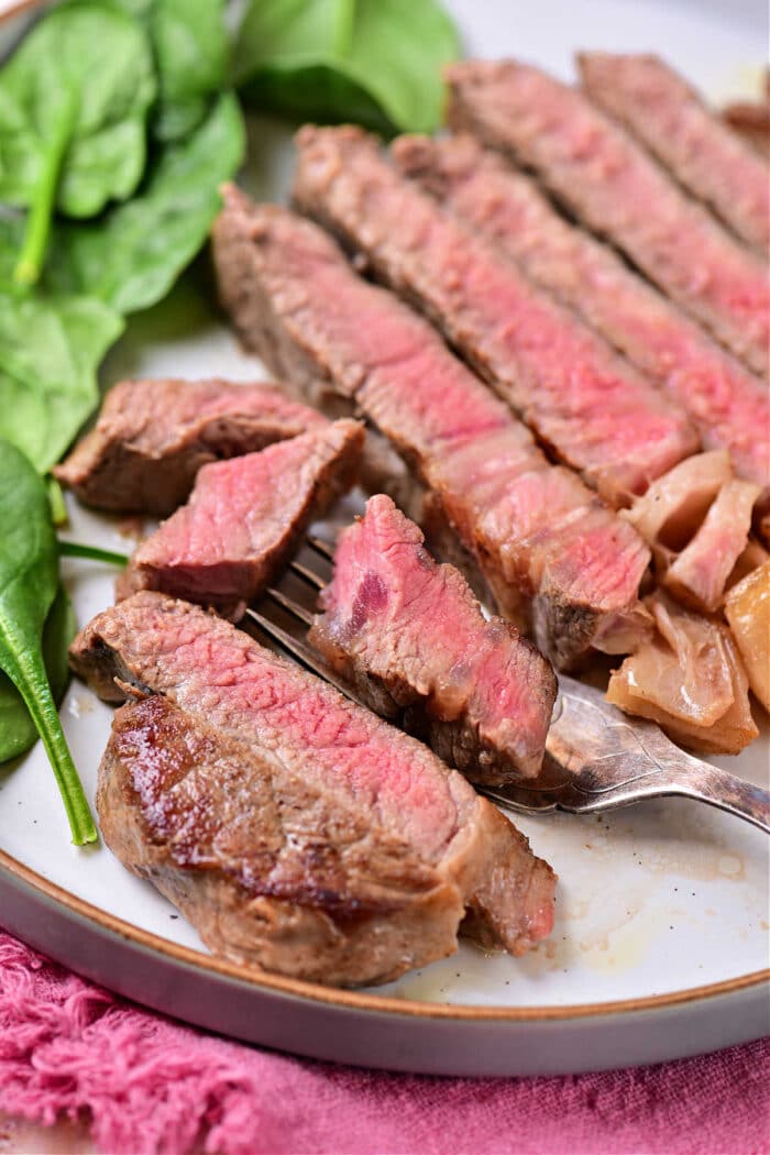 A plate featuring sliced medium-rare steak with visible pink centers is a perfect example of how to cook beef strip loin steaks. The steak is placed next to a leafy green, possibly spinach. A fork holds a piece of steak in front of the slices, with a pink cloth in the background adding to the presentation.