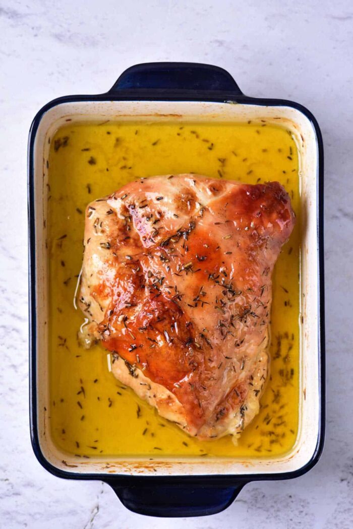 A cooked piece of turkey breast, prepared as Turkey Confit, is shown in a rectangular blue and white baking dish filled with golden liquid, likely broth or melted butter. The turkey is seasoned with herbs. The dish is placed on a light-textured surface.