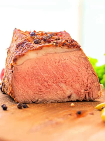 A close-up of a cooked roast beef, sliced to reveal its pink and tender interior. The beef's outer crust is seasoned, reminiscent of How to Cook a New York Strip Roast, and there are black peppercorns and mustard seeds on the wooden cutting board, along with some green leafy vegetables in the background.