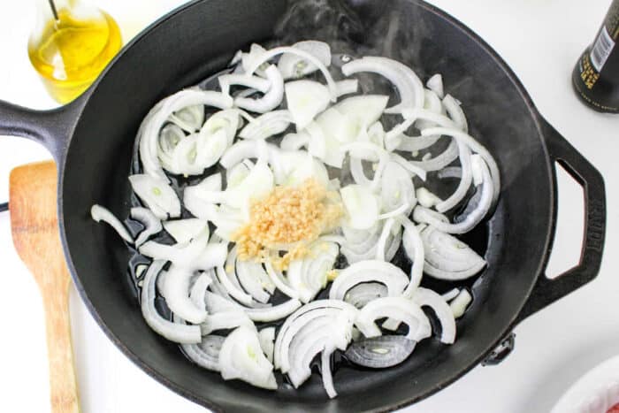 A cast iron skillet on a stovetop containing sliced onions and minced garlic, with steam rising from the pan. A wooden spatula is positioned next to the skillet, and a bottle of olive oil is visible in the background, setting the perfect stage for how to cook bratwurst to perfection.