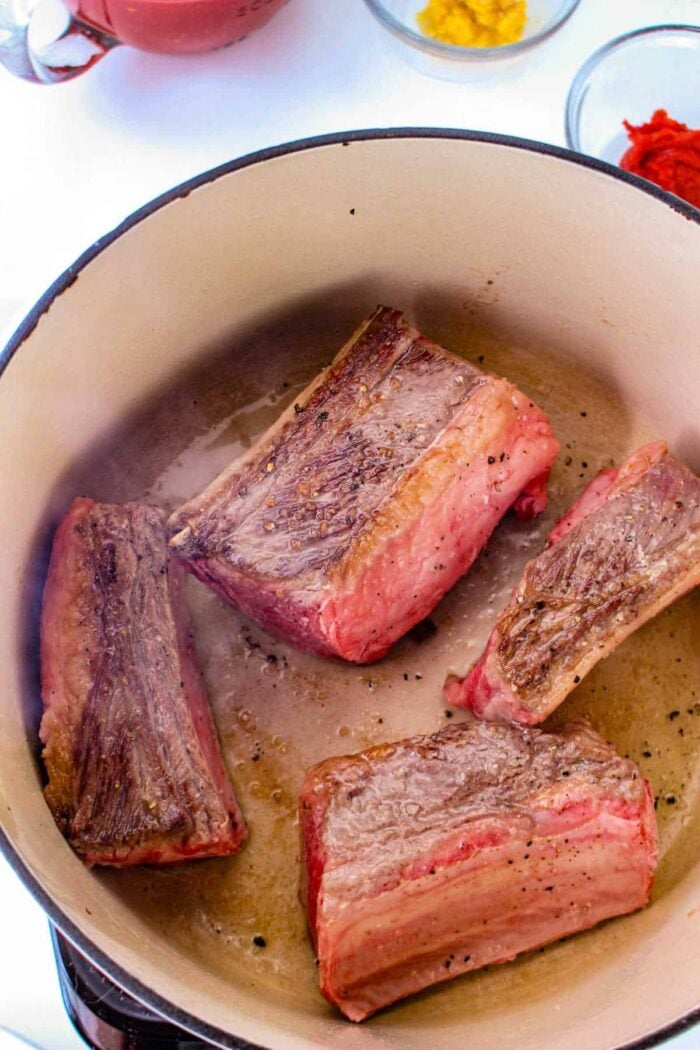 Four pieces of browned short rib meat are cooking in a large pot, perfect for tacos. The meat has a seared exterior. In the background, there are small bowls with ingredients, including a bright red and yellow substance. The pot and ingredients are set on a white surface.