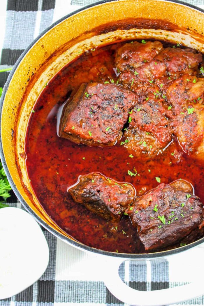 A top-down view of a pot filled with braised beef short ribs in a red sauce evokes the essence of short rib tacos. The dish is garnished with chopped herbs, and the pot rests on a checkered cloth. A small white dish is partially visible in the corner, completing this appetizing scene.