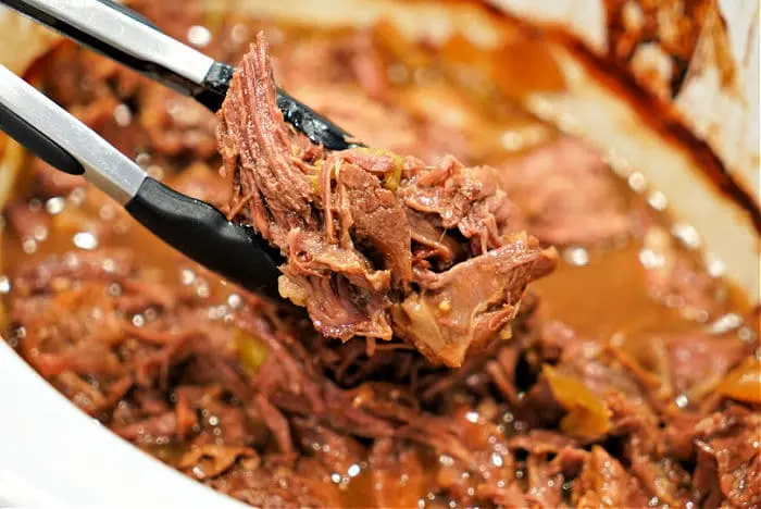 Close-up of a pair of tongs holding shredded beef chuck roast over a slow cooker filled with more tender, flavorful meat covered in rich juices.