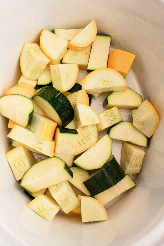 The image shows chopped pieces of zucchini and yellow squash inside a white container. The vegetables, perfect for a slow cooker recipe, are cut into uniform chunks, displaying their green and yellow peels.