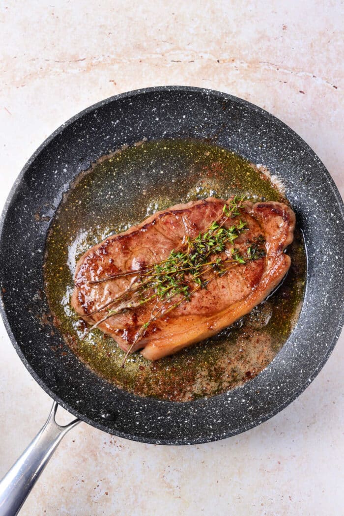 A cooked piece of steak garnished with sprigs of thyme rests in a black non-stick frying pan on a beige countertop. The steak sits in a small pool of rendered fat or sauce, providing a mouthwatering glimpse into how to cook beef strip loin steaks to perfection.