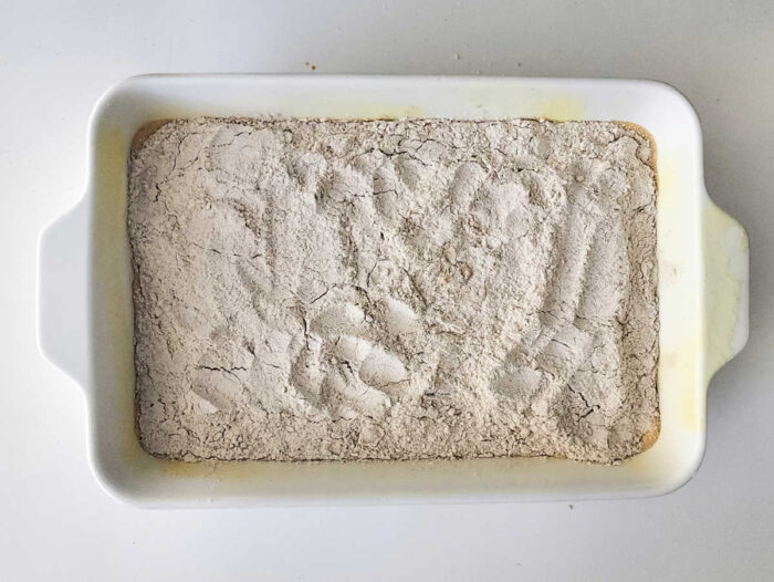 A white baking dish with a layer of flour and handprints visible on the surface, ready to become the base for a delicious Sweet Potato Dump Cake.