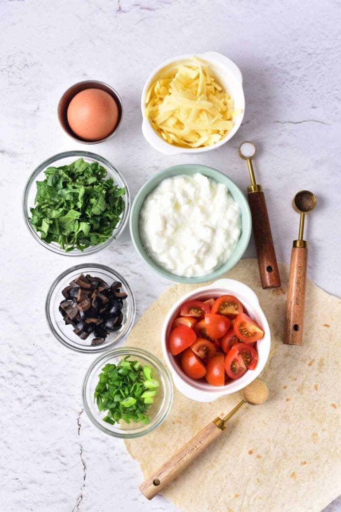 A flat lay image showcases various ingredients for a meal, perfect for making Cottage Cheese Quiche. These include grated cheese, an egg, chopped spinach, ricotta or cottage cheese, cherry tomatoes, chopped mushrooms, green onions, and a tortilla. Two wooden-handled scoops are placed nearby.