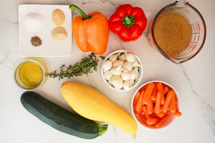 An assortment of ingredients for slow cooker vegetables are laid out on a light-colored surface. These include a yellow squash, zucchini, baby carrots, a small bowl of garlic cloves, red and orange bell peppers, fresh thyme, olive oil, seasoning mix, and a measuring cup with broth.