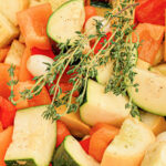 A close-up of slow cooker vegetables including zucchini, red bell peppers, and onions being cooked in a pot. The vegetables are topped with sprigs of fresh thyme. A red spatula is placed within the mixture.