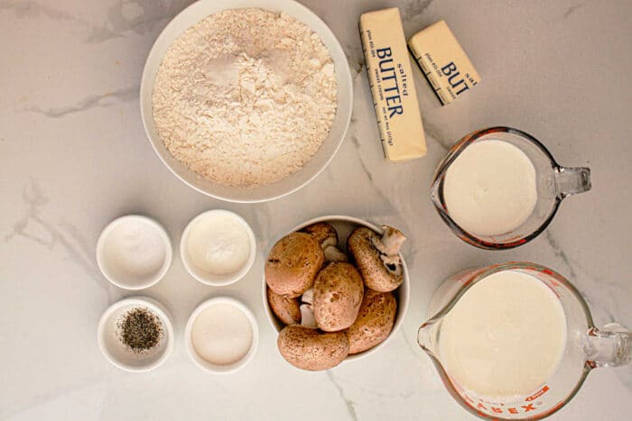 A top-down view of cooking ingredients on a white marble surface. There are flour, two sticks of butter, milk, cream, whole mushrooms, and small bowls containing salt, pepper, and sugar—perfect essentials for preparing Slow Cooker Biscuits and Gravy.