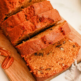 A loaf of sweet potato bread partially sliced on a wooden cutting board. The bread is golden brown with visible specks of nuts. Whole pecans and a cinnamon stick are placed beside the board on a white surface. A gray cloth is partially visible in the corner.