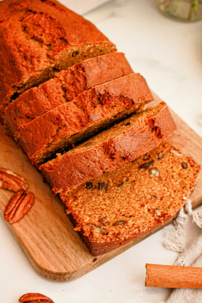 A sliced loaf of banana nut bread sits on a wooden cutting board, with some pecan halves scattered around. The bread has a golden-brown crust and visible pecan pieces in each slice, reminiscent of Sweet Potato Bread with Cake Mix. A small piece of cloth and a cinnamon stick are partially visible beside the board.