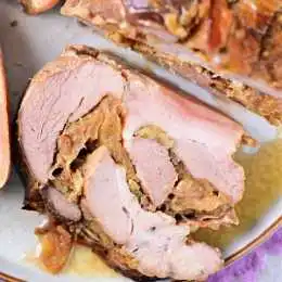 A close-up shot of a sliced roast, showing layers of tender meat and a stuffed filling inside. The roast is served on a white plate with a light brown rim. The filling appears to be vegetables and possibly spices, with a bit of gravy pooling around—perfect for delicious family meals.