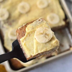 A close-up of a spatula holding a square slice of banana cake topped with banana slices. The rest of the banana bread squares with the same topping are visible in the background inside a baking pan.