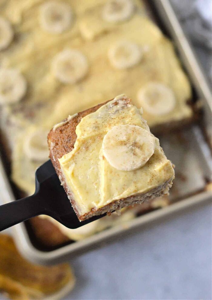 A close-up of a spatula holding a slice of banana bread topped with a spread and a banana slice. In the background, there is a baking tray containing more pieces of similarly topped treats from an easy banana cake recipe with cake mix.