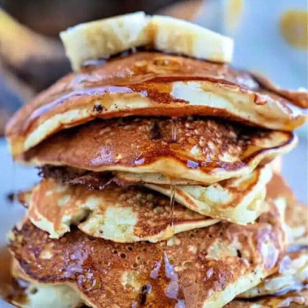 A stack of fluffy Banana Chocolate Chip Pancakes topped with banana slices and drizzled with maple syrup on a white plate.