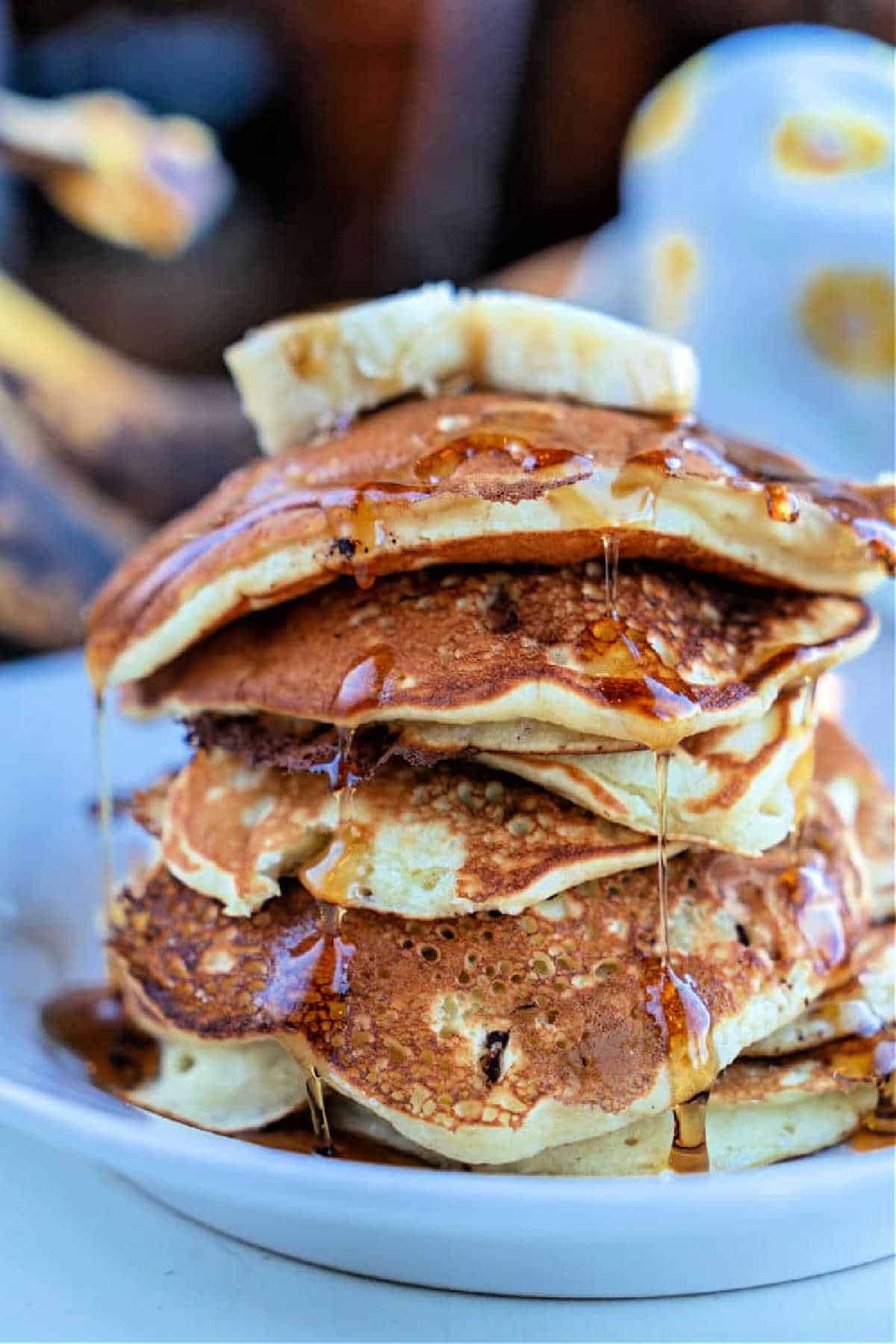 Stack of banana chocolate chip pancakes topped with banana slices and drizzled with syrup on a white plate.