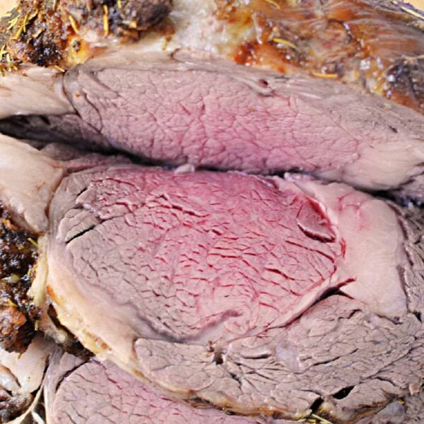 A close-up of a sliced, medium-rare Boneless Rib Roast. The beef is browned on the outside and pink with a slightly red center on the inside. It is garnished with sprigs of fresh herbs in the background. The meat is placed on a wooden cutting board.