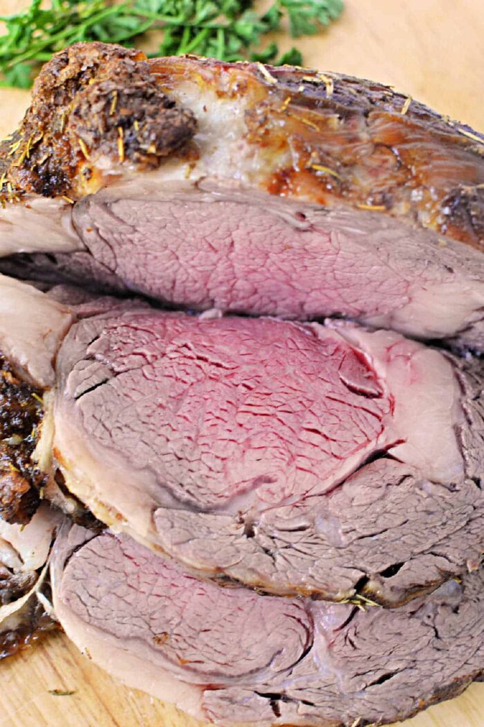 A close-up of a sliced, medium-rare Boneless Rib Roast. The beef is browned on the outside and pink with a slightly red center on the inside. It is garnished with sprigs of fresh herbs in the background. The meat is placed on a wooden cutting board.