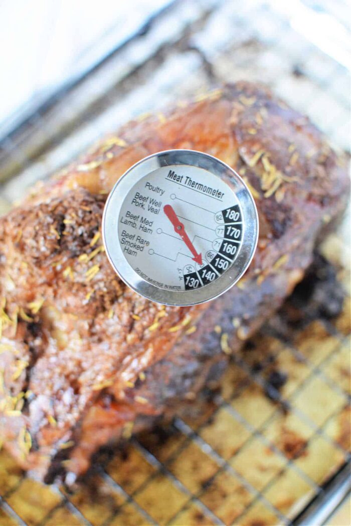 A meat thermometer is inserted into a cooked boneless rib roast, showing a temperature reading. The roast, seasoned with herbs, rests on a wire rack set on a baking sheet. The thermometer's dial displays various meat types and corresponding temperature ranges.