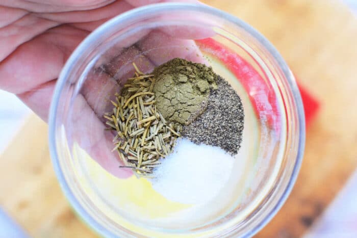 A hand holding a clear glass bowl containing a mix of dried rosemary, ground black pepper, salt, and another greenish-brown spice. Perfect for seasoning your Boneless Rib Roast Recipe. A wooden cutting board is visible in the background.